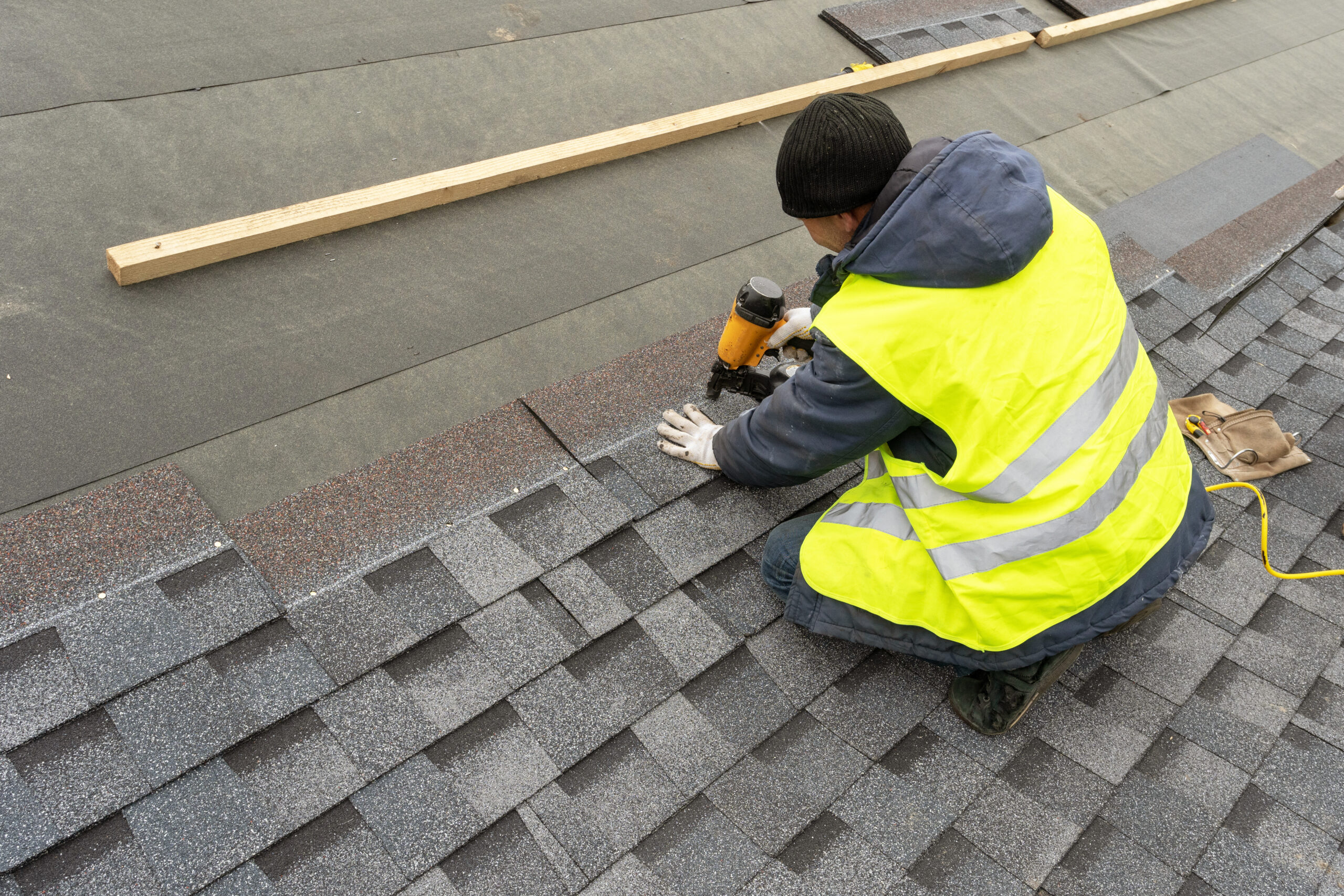 Working man is using a nail gun to install asphalt shingles on a roof